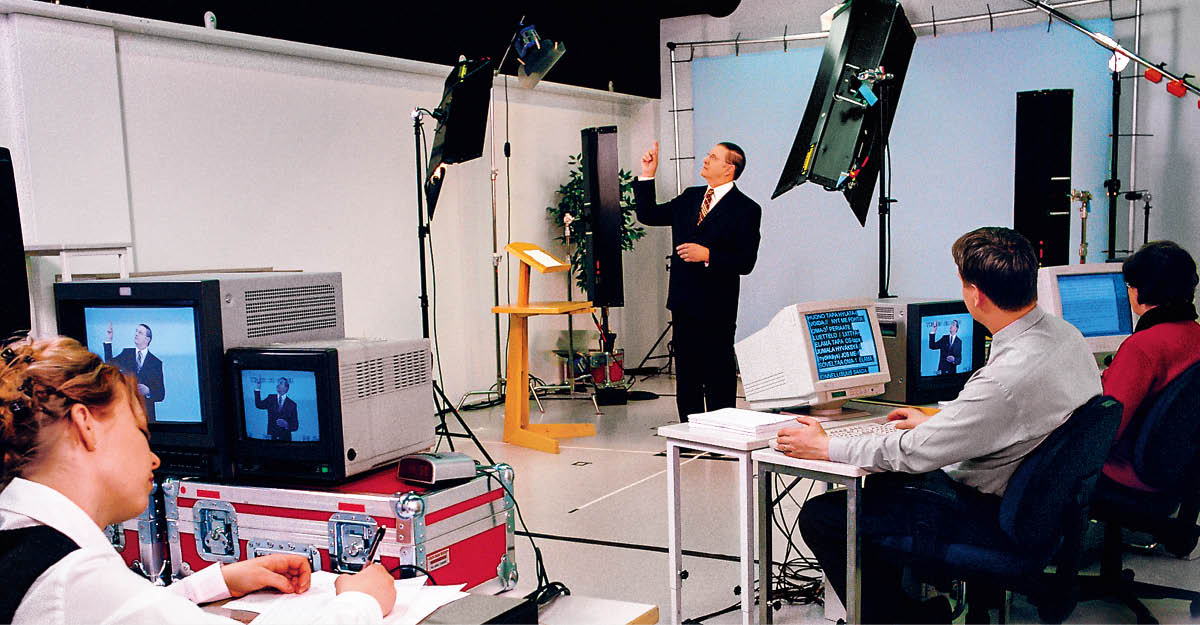 Tapani gebärdet im Studio für eine Videoaufnahme. Drei andere, darunter auch Maire, verfolgen die Aufnahme auf Computer­bildschirmen.