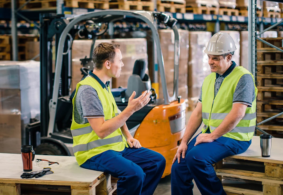 Un hermano entregando una invitación a la Conmemoración a un familiar que no es Testigo mientras están sentados en la cocina. Sus esposas hablando y cocinando detrás de ellos