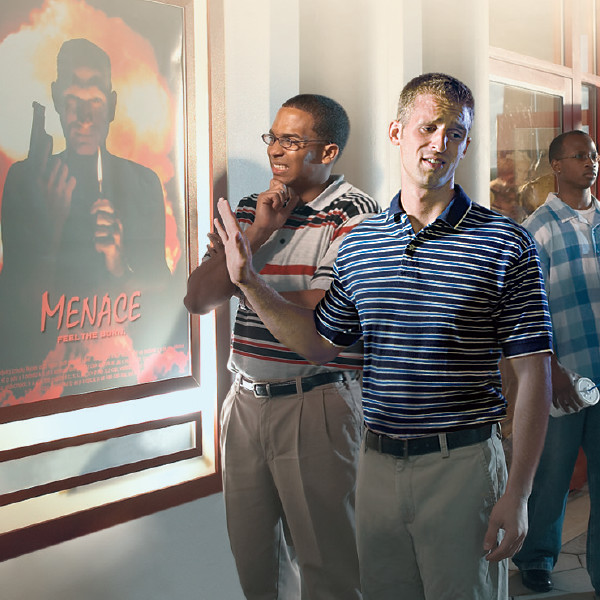 Two brothers standing in front of a poster that advertizes a violent movie. One brother grimaces, and the other throws his hand in the air and turns away.