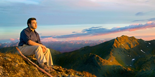 A man sits and looks out at a mountain range and beautiful sky