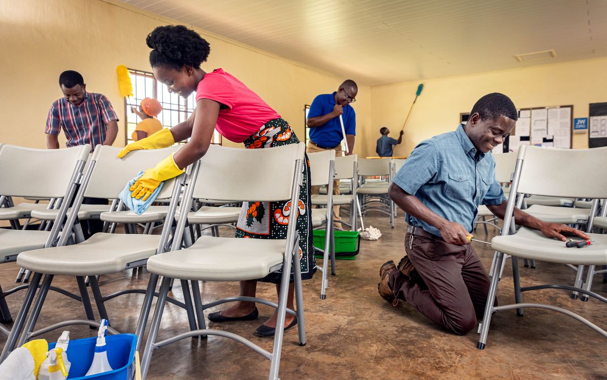 Brothers and sisters thoroughly cleaning a Kingdom Hall while another brother repairs one of the chairs.