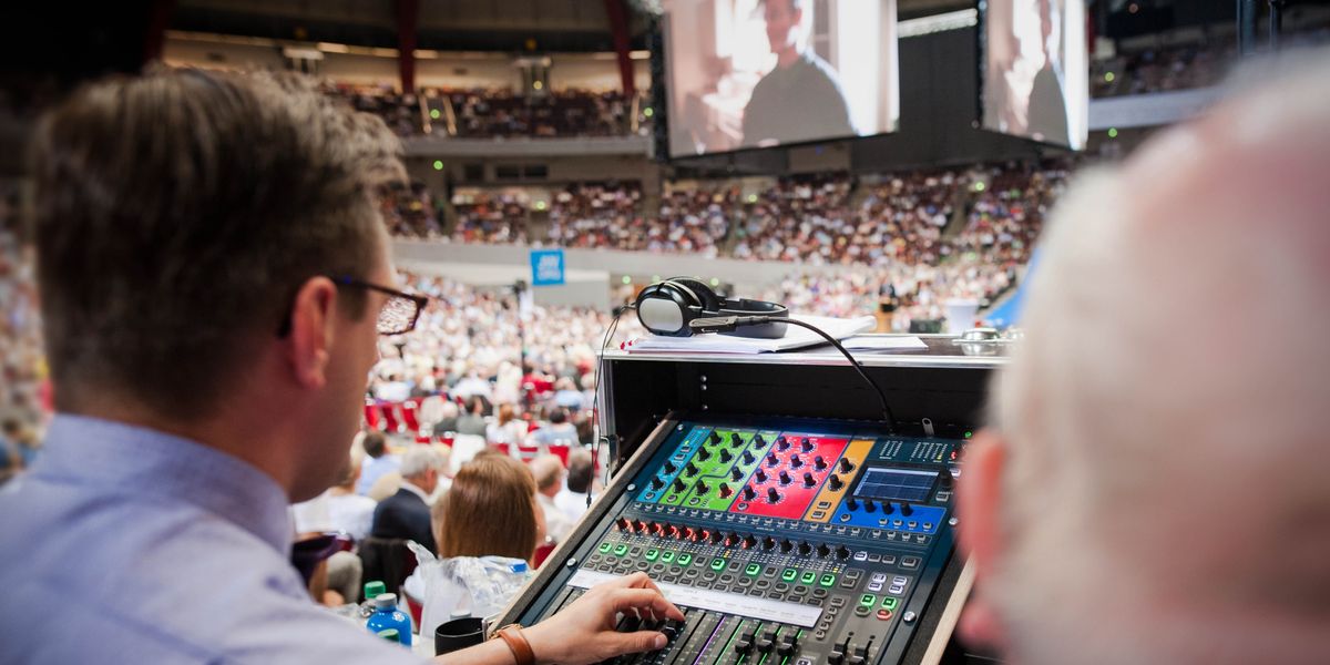 A brother adjusting audio levels at a convention while the audience watches a video on large screens.