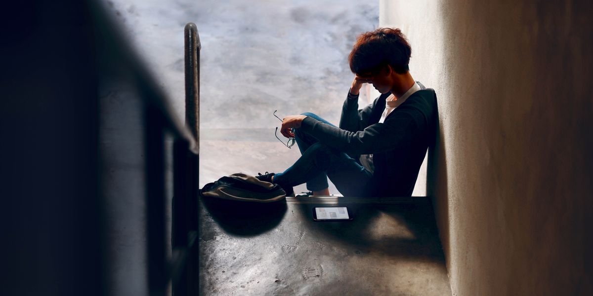 A teenager resting his head in his hand as he sits alone at the bottom of a set of stairs in a dark hallway.