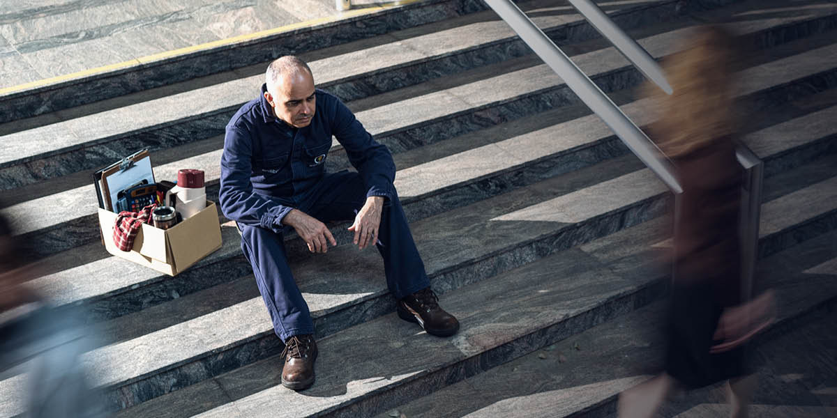 A despondent man sitting on the steps outside his workplace. Next to him is a box of personal belongings from his job.