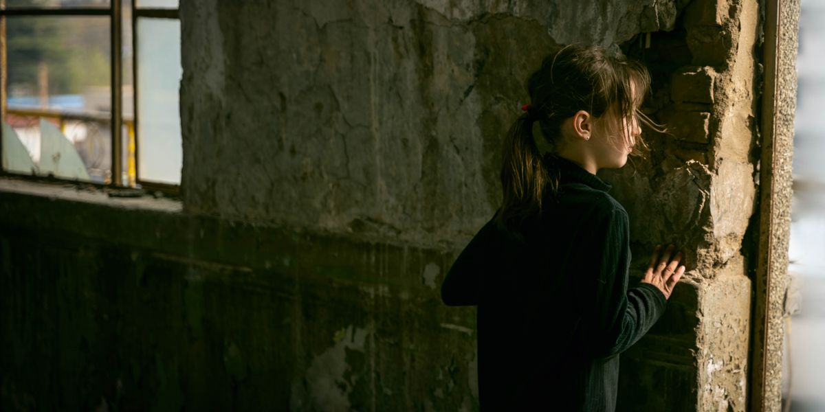 A young girl peering out of an opening in a wall of a destroyed building.