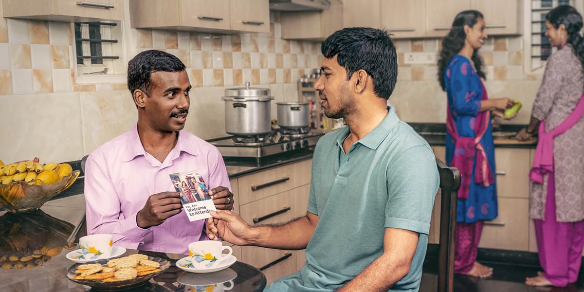 A brother giving a non-Witness relative an invitation to the Memorial while they sit at a kitchen table. Their wives talking and preparing a meal in the background.