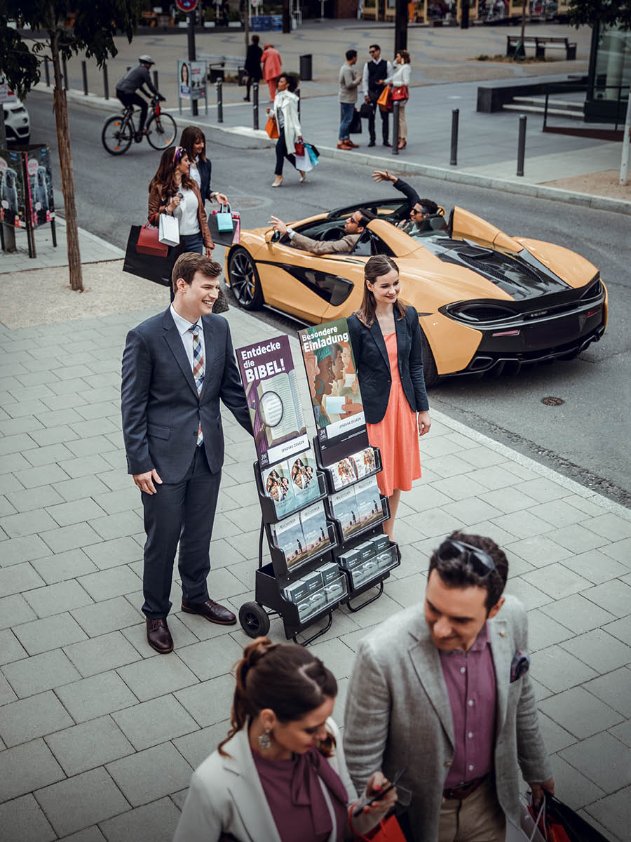 A couple happily engaging in public witnessing while passersby are shopping and driving expensive cars.