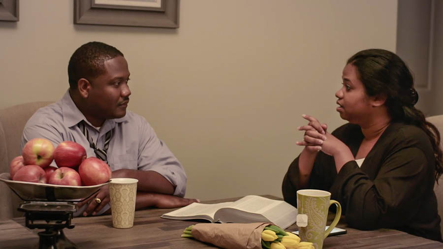 A scene from the video ‘We Must ‘Run With Endurance’​—Obey the Rules of the Contest.’ Brother and Sister Calou calmly discuss their challenges. An open Bible is on the table.