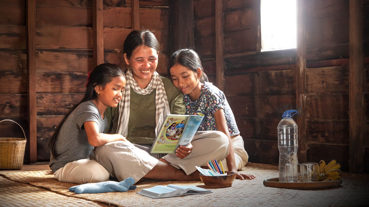 A mother and her two daughters sitting on the floor of their modest home, joyfully reading the book “Learn From the Great Teacher.”