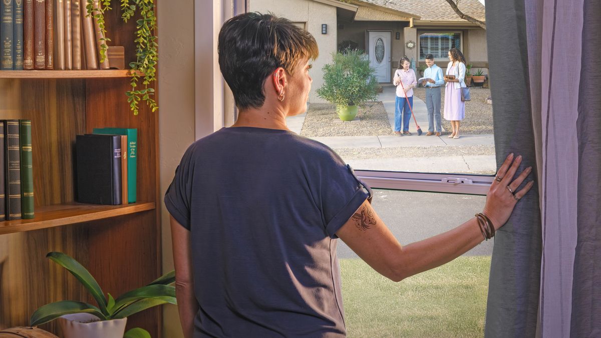 A woman looking out her window as a couple of Witnesses preach to her neighbor. She has older editions of the “New World Translation” and a songbook on her bookshelf.