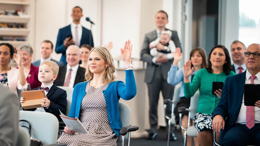 A mother sits with her little boy during a congregation meeting and raises her hand to participate, while the father holds their baby girl in the back of the auditorium