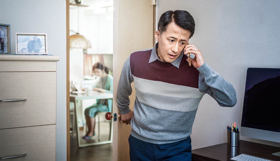 An elder talking on the phone and shutting the door behind him while his wife studies in the other room.