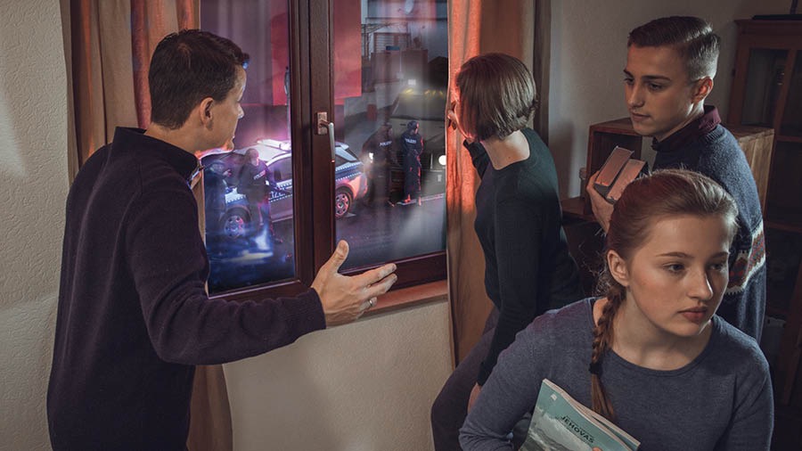 The family depicted earlier. The parents look out a window at the police who are outside their home while the son and daughter calmly prepare to hide Bible-based publications.