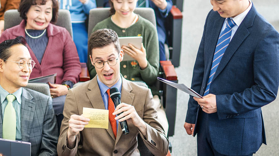 A meeting in the Kingdom Hall where a brother who is learning a foreign language reads a handwritten comment. Others in attendance look on with respect.