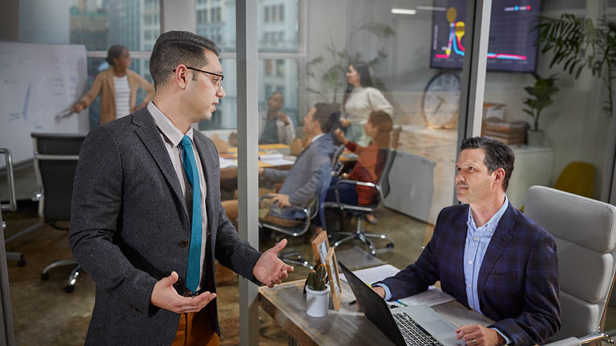 A brother explaining to his employer why he cannot work overtime. In the background, other employees meet in a conference room.