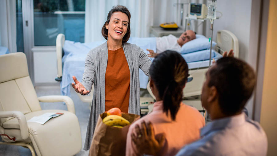 A sister warmly welcomes a couple who have come to visit her husband in the hospital. The couple have brought some food as a gift.