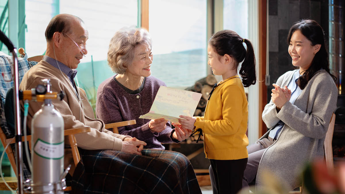 A mother and her young daughter giving a card the daughter has made to an elderly couple. The elderly brother uses an oxygen tank to help him breathe.
