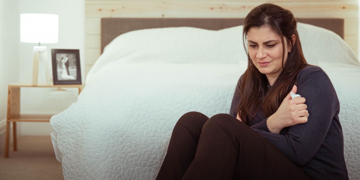 A sister crying as she sits on the floor next to her bed. A framed wedding picture is in the background.