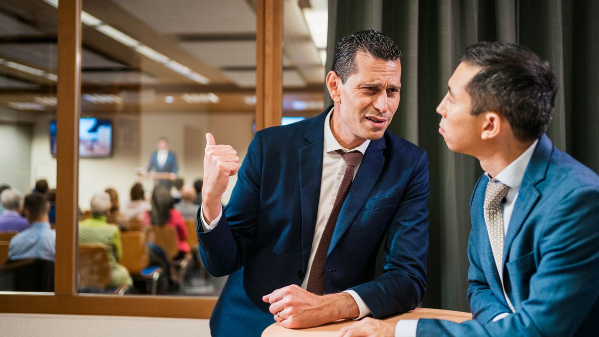 Two brothers conversing with each other during a congregation meeting. One of the brothers frowns and gestures toward a brother who is giving a talk at the meeting.