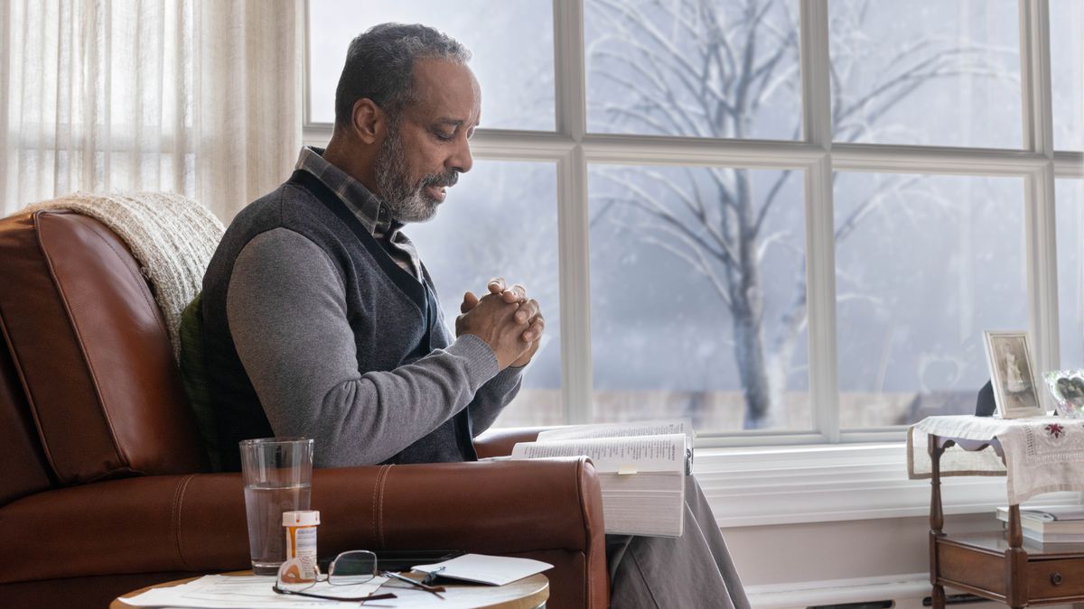 An elderly brother sitting in his home and praying in the wintertime. An open Bible is on his lap, and a bottle of medication is on a table next to him.