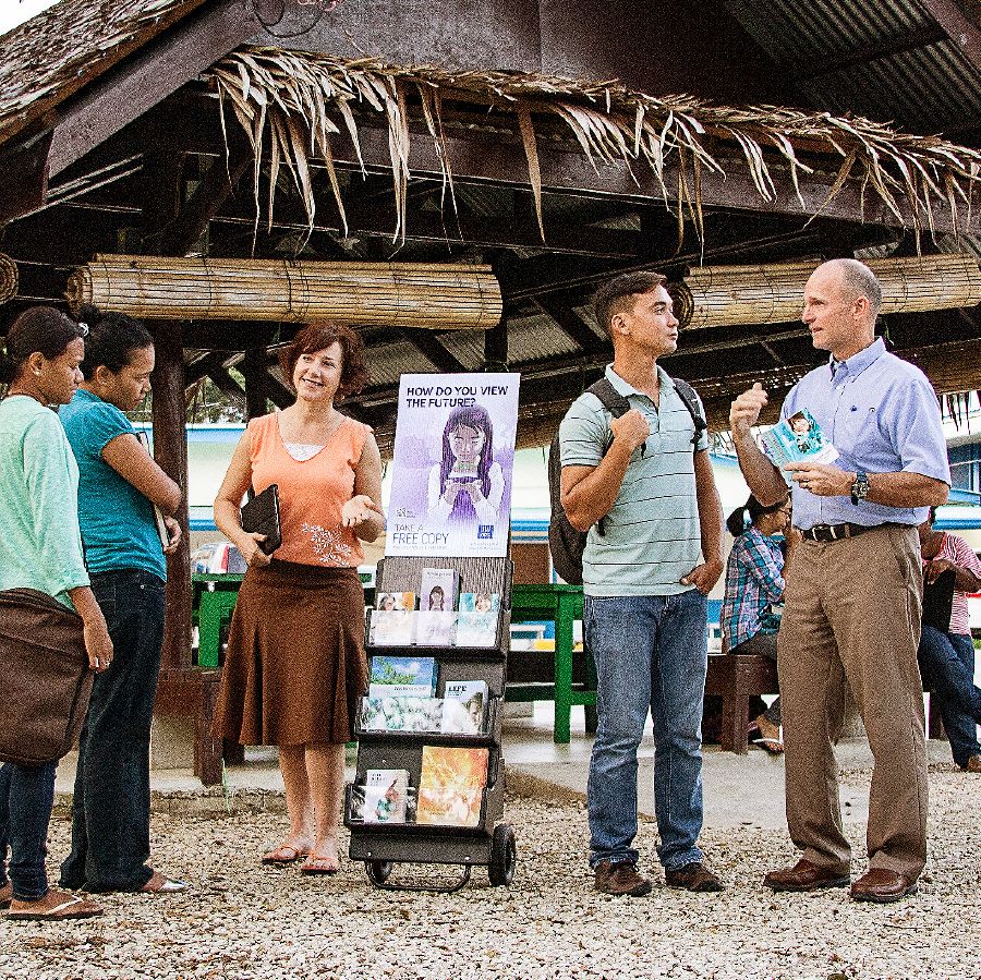 Missionaries Roxanne and Brian stand beside a literature cart near a college campus in Palau and converse with students