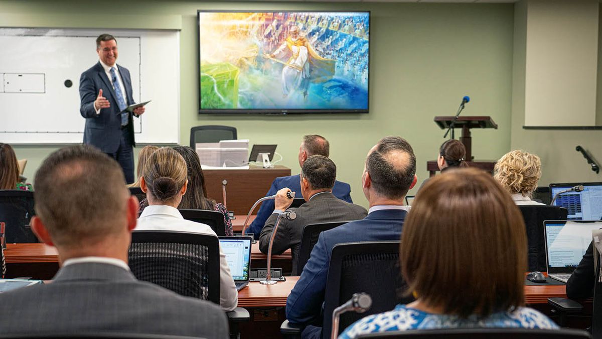 Un profesor dando clases. Detrás de él, hay una pantalla en la que aparece Jesús entregándole el Reino a Jehová Dios. Los estudiantes usan los micrófonos de sus escritorios para comentar.