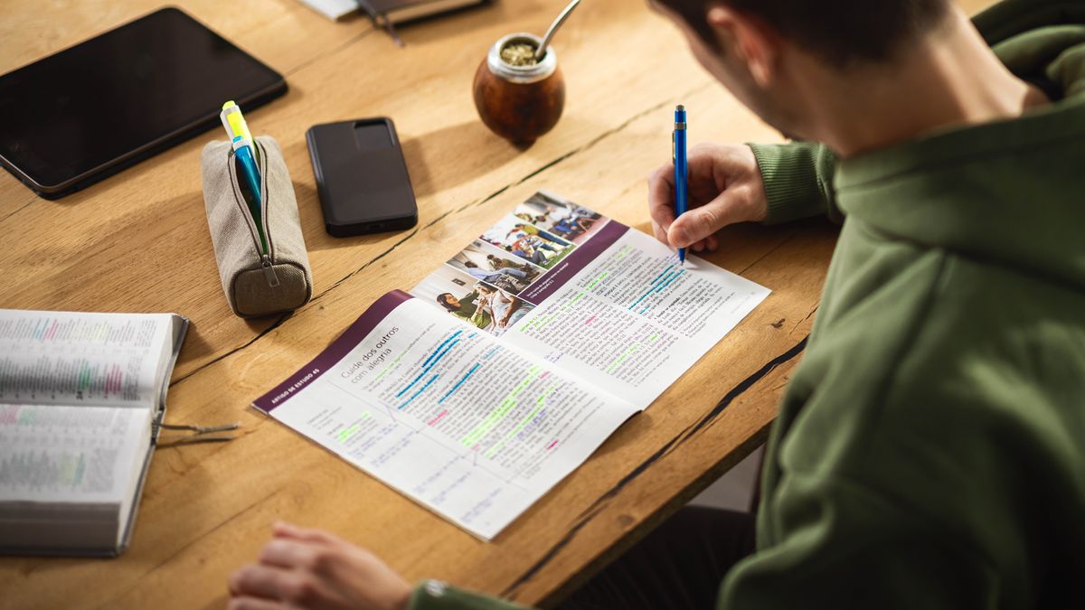 Un hermano en su escritorio estudiando y marcando las respuestas en una “Atalaya” de estudio en papel. También tiene una Biblia impresa con varios versículos marcados.