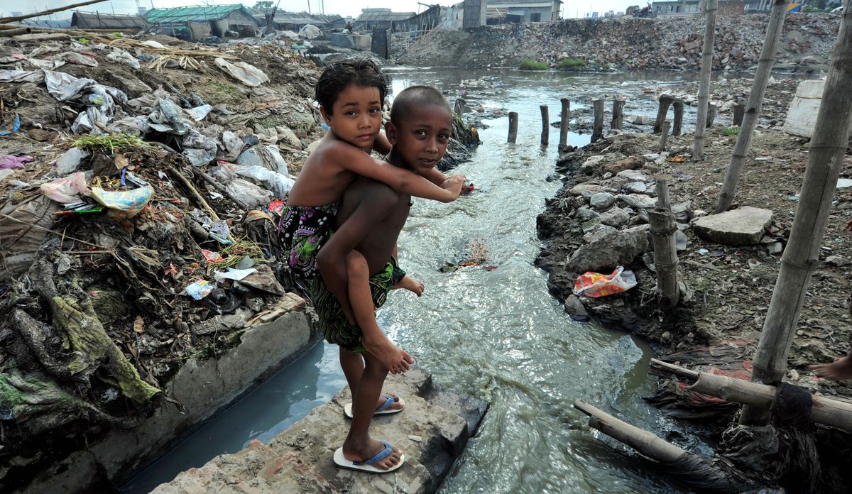En la orilla de un río muy contaminado, un niño carga en la espalda a un niño más pequeño.