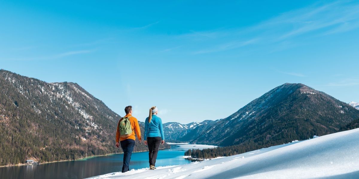 Unha parella camiñando por unha costa cuberta de neve e mirando cara á un lago que está rodeado de montañas cheas de árbores. O ceo está azul e despexado.
