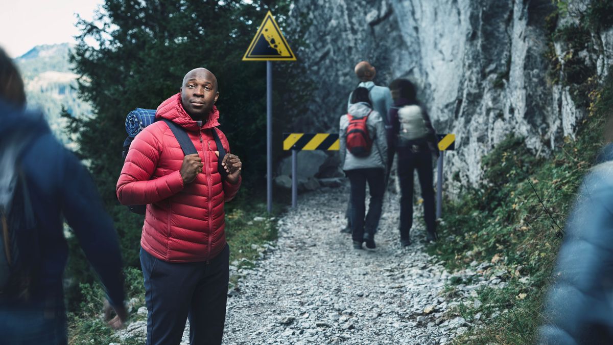 Jèn gason an ki kanpe nan yon wout an wòch bò yon pankat ki gen siy pou di fè atansyon. Lòt moun k ap monte mòn nan pa okipe pankat la.