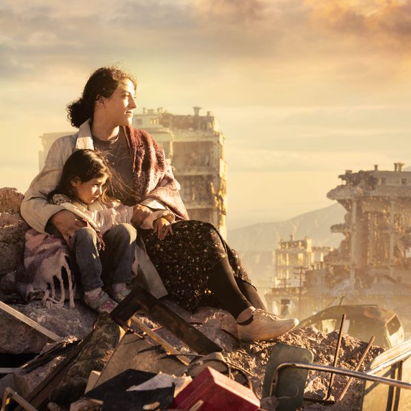 A woman with her arm around her young daughter while they sit on a pile of rubble in a city that has been devastated by war. They are surrounded by destroyed buildings.