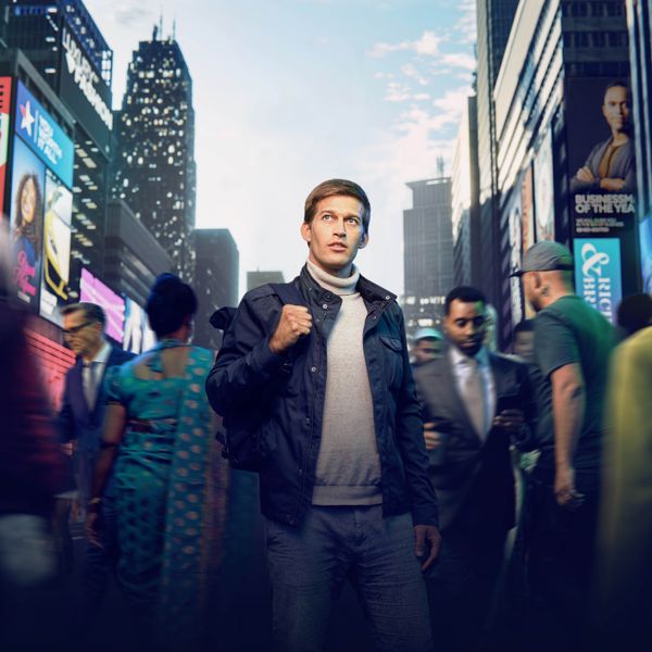 Un giovane uomo in piedi in una strada piena di gente. Persone di diversa provenienza gli passano accanto. Lui ha lo sguardo rivolto verso l’alto, come se stesse cercando di capire dove andare.