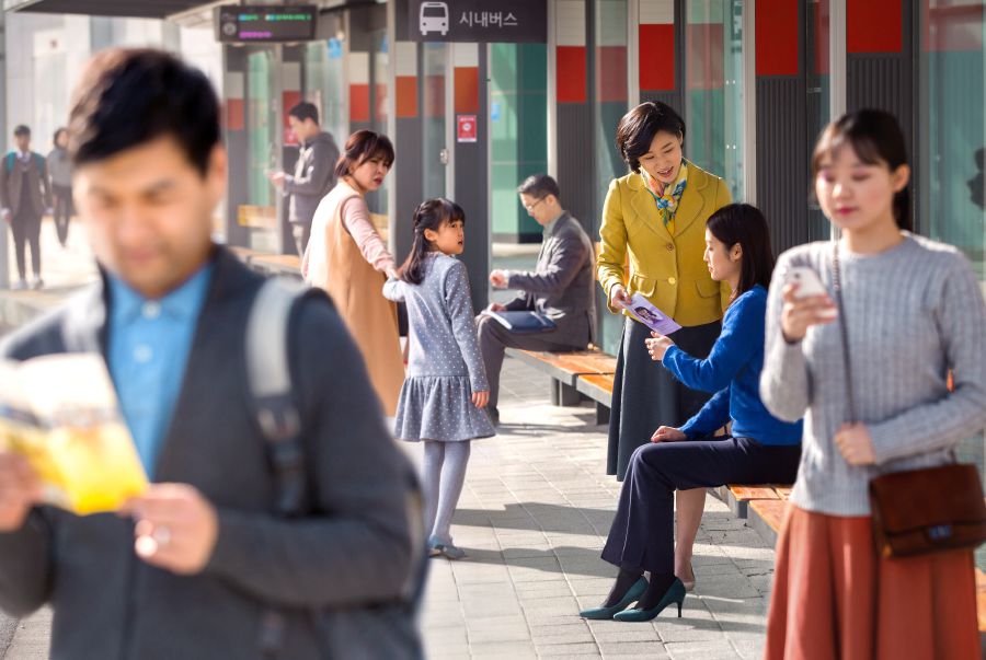 한 자매가 버스 정류장에서 한 여자에게 전도지를 전하고 있고 주위에 있는 많은 사람들은 관심을 나타내지 않는 모습
