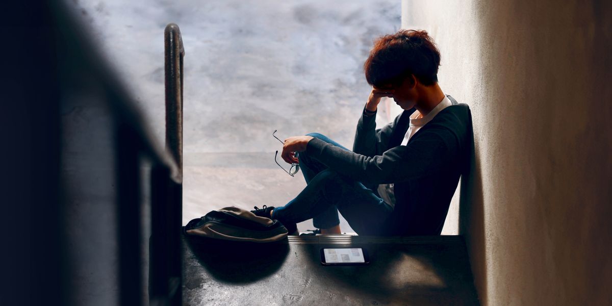 The young man sitting down by his self in the dark hall way holding his head.