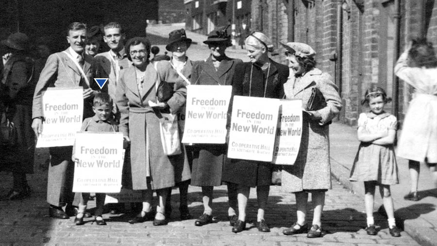 Stephen Hardy as a young boy, preaching with his parents and several others. They hold posters advertising the title of the public talk at an assembly.