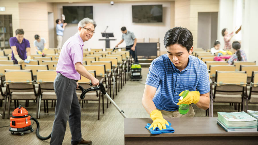 The young brother helping to clean the Kingdom Hall. The elder who earlier gave him advice observes from a distance.