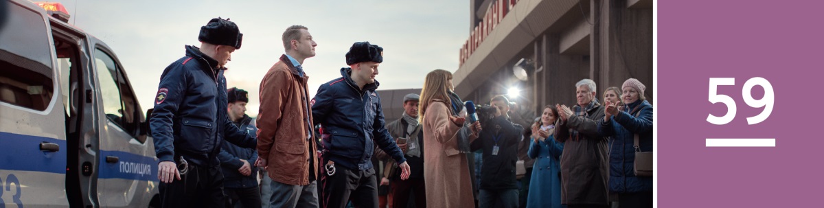 59. nodarbība. Divi policisti ved uz tiesu rokudzelžos saslēgtu Jehovas liecinieku. Pie ieejas stāv reportieri un Jehovas liecinieki, kas atnākuši atbalstīt savu ticības biedru.