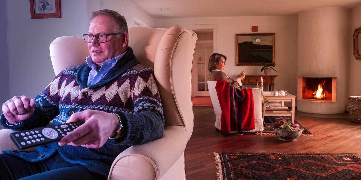 En mann sitter og ser på tv mens kona sitter i andre enden av stua og strikker
