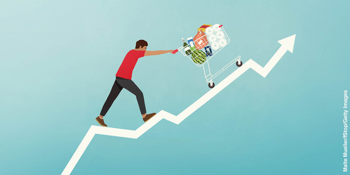 Gráfico com uma linha mostrando o grande aumento nos preços. Um homem está empurrando um carrinho cheio de compras, subindo até o fim da linha do gráfico.