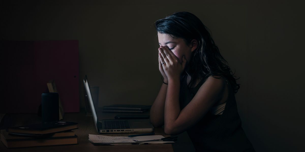 Uma adolescente triste. Ela está sentada tarde da noite em frente ao seu computador e está cobrindo o seu rosto com as mãos.