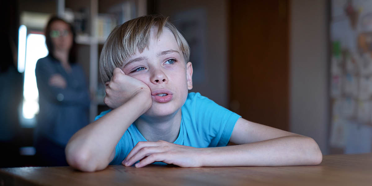 Мама смотрит на сына, который со скучающим видом сидит за столом и смотрит в окно.