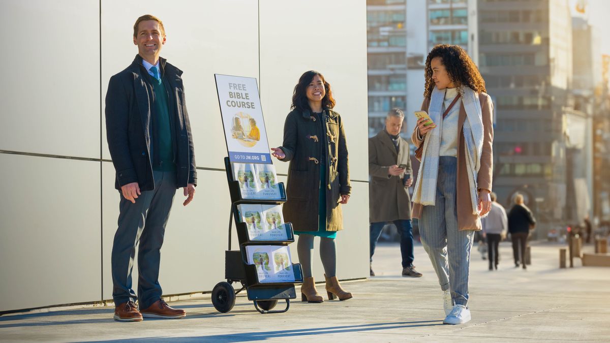 Муж и жена проповедуют со стендом для литературы, на котором написано: «Бесплатный библейский курс»; проходящая мимо женщина смотрит на стенд.