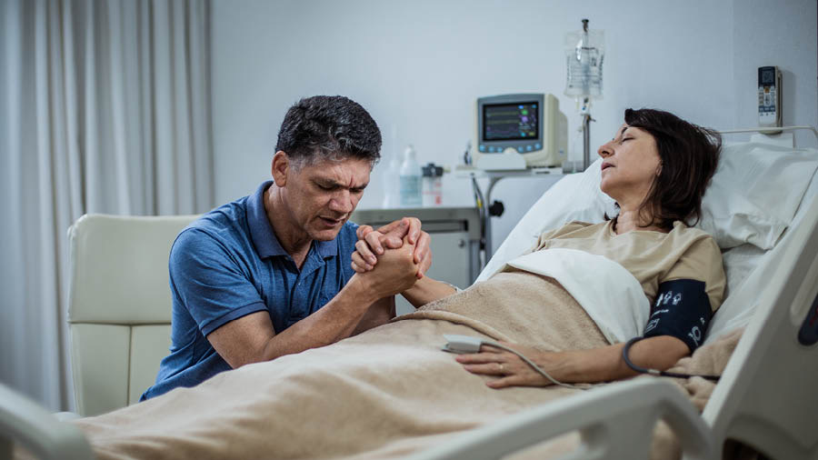 Муж се моли са својом женом док она лежи на болничком кревету.