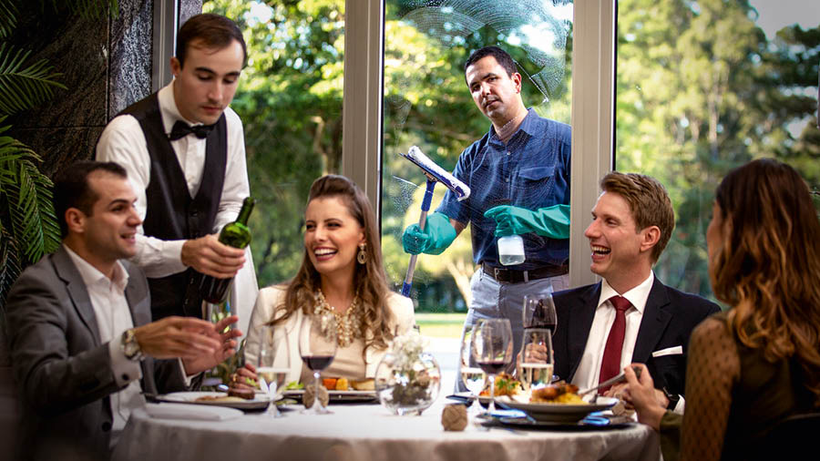 Två par skrattar och äter tillsammans på en exklusiv restaurang. En broder putsar fönster på restaurangen och tittar avundsjukt på dem.